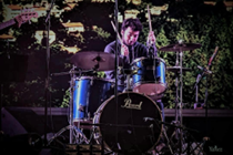 A drummer performing on stage, focused on playing a blue drum set with a backdrop of greenery.