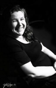 A smiling woman with curly hair sits casually, illuminated in a black and white portrait.