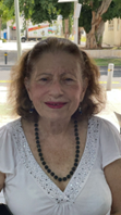 A smiling older woman with curly hair, wearing a white top and a necklace, is seated outdoors.