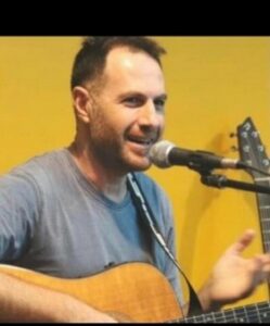 A man with short hair is singing into a microphone while playing an acoustic guitar, set against a yellow background.